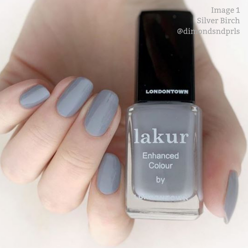 Close-up of a hand with painted nails holding a bottle of LONDONTOWN Lakur Enhanced Colour nail polish in the shade "Silver Birch." The nails, painted in a matching grey color, showcase this cruelty-free and vegan beauty marvel.