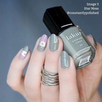 Close-up of a hand with grey and lavender manicured nails, holding a bottle of LONDONTOWN Star Moss (Professional) vegan nail polish in an olive shade. The Instagram handle @constantlypolished is also in the image.
