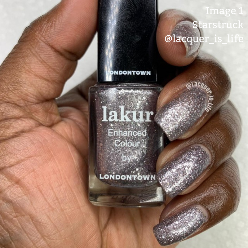 Close-up of a hand holding a bottle of LONDONTOWN Starstruck Lakur nail polish, with the nails painted in a matching glittery shade. This shimmer polish is both vegan and cruelty-free. The Instagram handle @lacquer_is_life is printed on the hand.