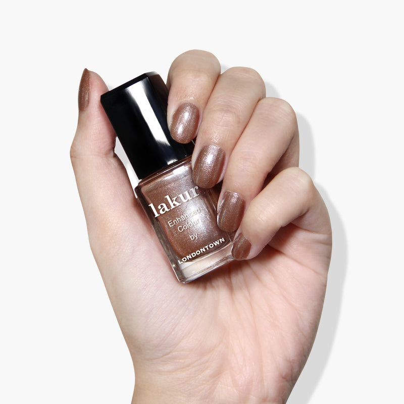 A hand with manicured nails is holding a bottle of vegan, cruelty-free LONDONTOWN Lakur nail polish in the Sugarcane shade, which is also visible on the nails.