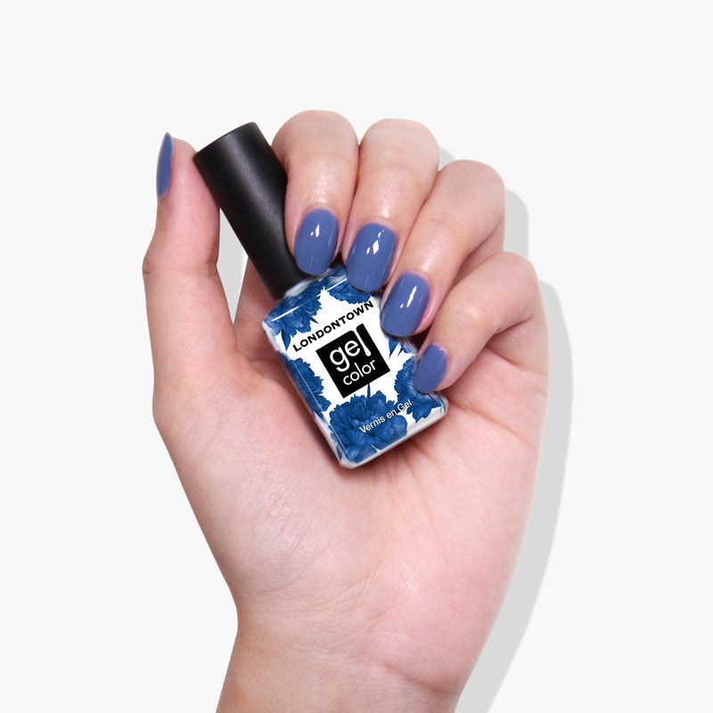 A hand holds a bottle of LONDONTOWN Gel Color nail polish in the shade "To the Moon," showcasing nails painted in a matching crème blue hue.