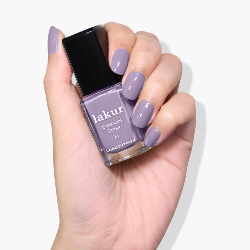 A hand holding a bottle of "Trolley Away" nail polish by LONDONTOWN, with nails painted to match. This vegan and cruelty-free nail polish boasts a stunning light purple shade.