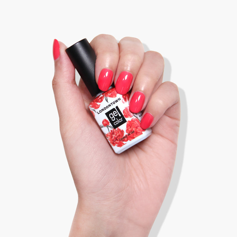 A hand with manicured nails holds a bottle of LONDONTOWN's Weekend Cheers coral pink crème gel nail polish with a floral design on the label.