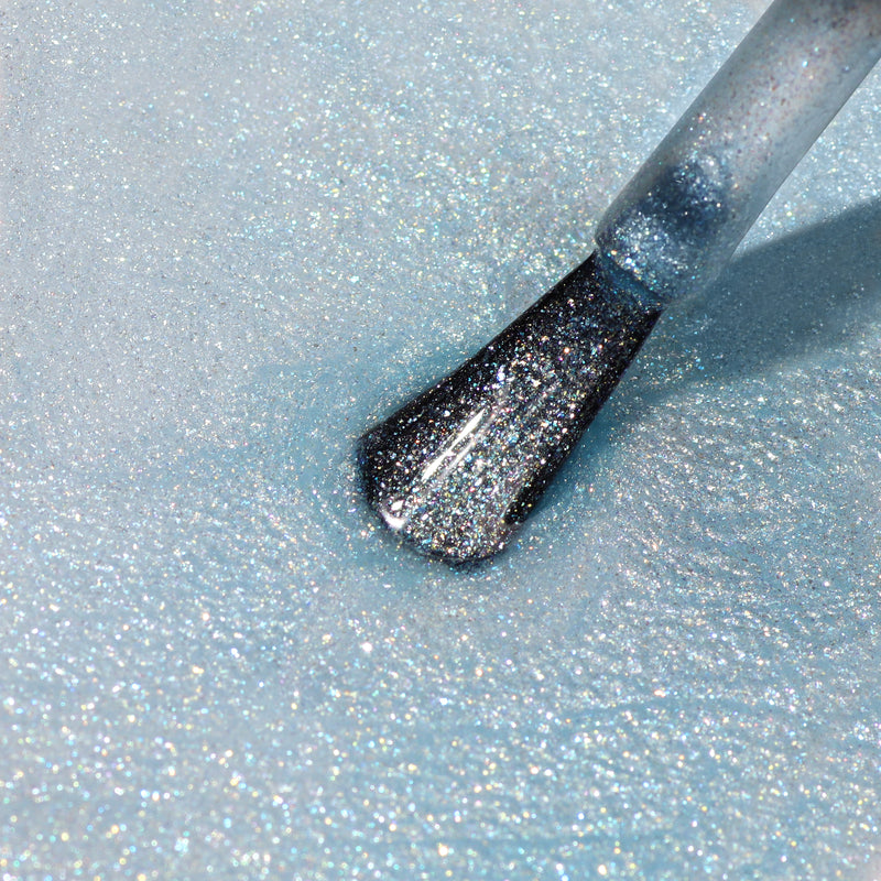 Close-up of a LONDONTOWN nail polish brush applying Whipped Blueberry, an ultrafine glitter nail polish, on a sky blue, sparkly surface.