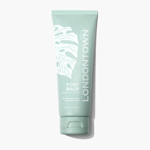 A tube of LONDONTOWN Foot Balm, featuring a green, leafy design and white text that highlights its benefits for smoother soles and softer heels. Infused with eucalyptus and lemongrass for a refreshing touch, this balm is available in a 4 fl oz (120 ml) size.