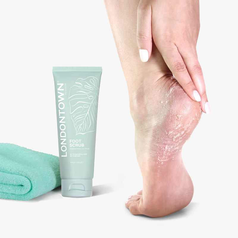 A hand applying LONDONTOWN Foot Scrub, enriched with natural sugar crystals and essential oils, to the heel, with a tube labeled "LONDONTOWN Foot Scrub" and a folded turquoise towel placed next to it.